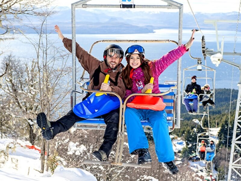 Personas descendiendo por una colina de nieve en trineos o culipatines, con árboles y cielo nublado de fondo.